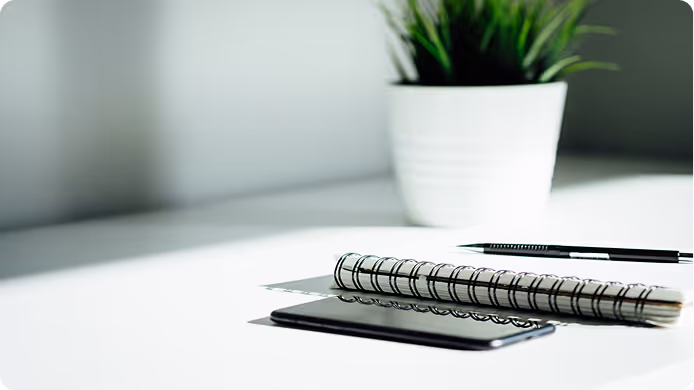 A grayscale photograph of a desk with a notebook, pen, smartphone, and a potted plant.