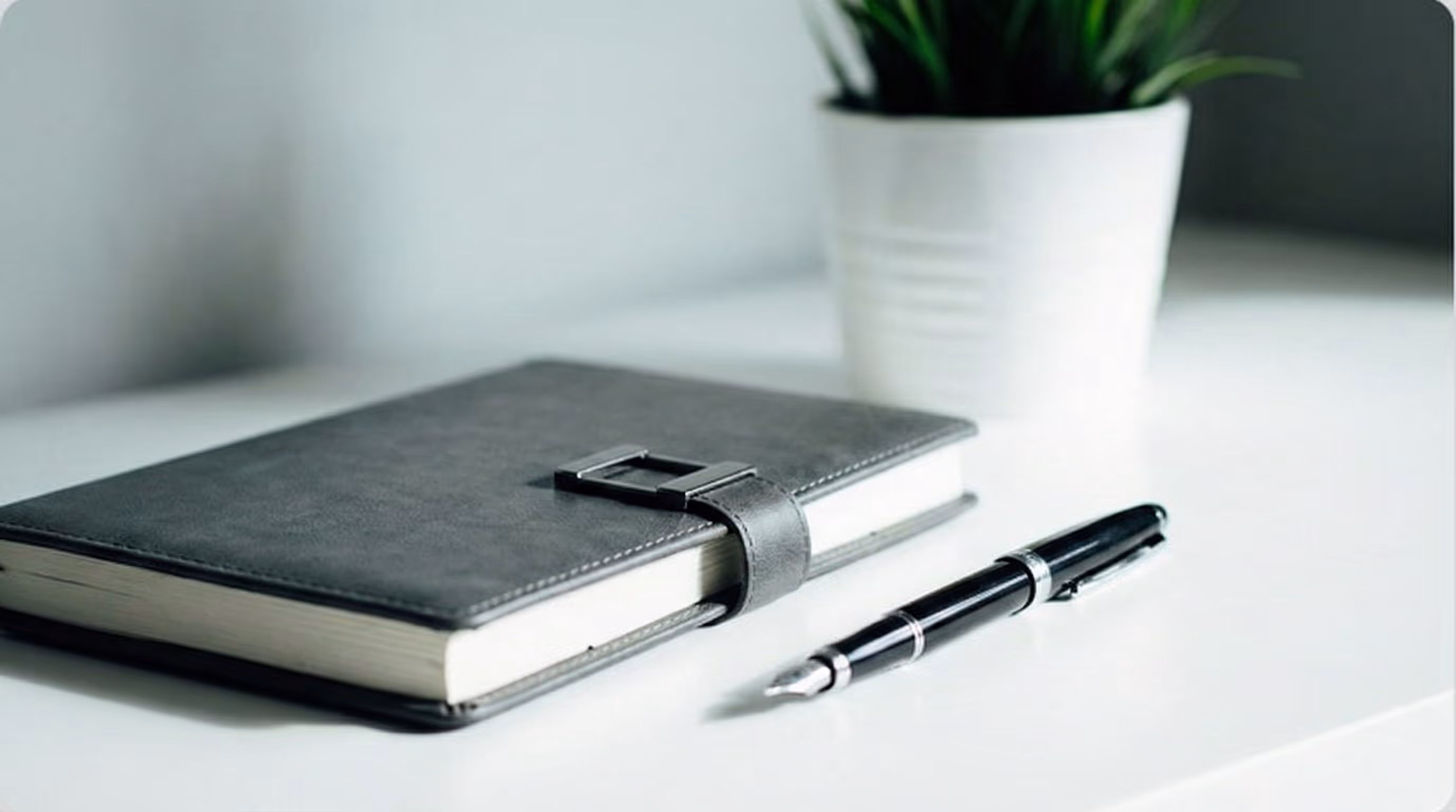 A grayscale photograph of a desk with a notebook, pen, smartphone, and a potted plant.